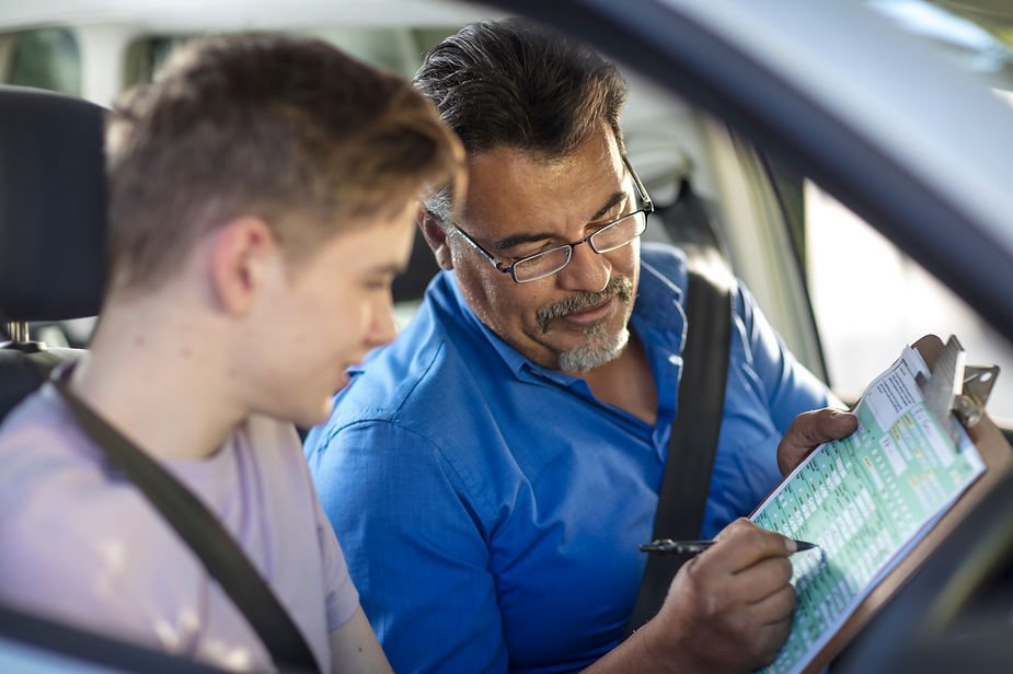 Man in blue shirt marks checklist, seated in a car with a young male driver. Bright, focused atmosphere. Clipboard and pen visible.