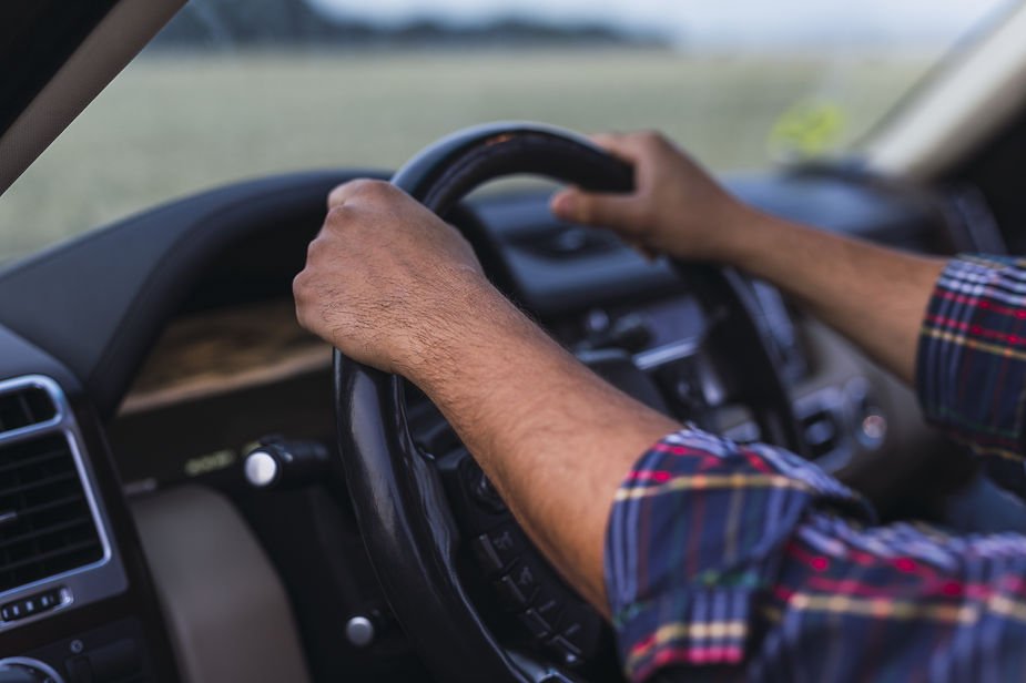 Hands on a black steering wheel inside a car. The person wears a plaid shirt. Background shows a blurred view of fields through the window.