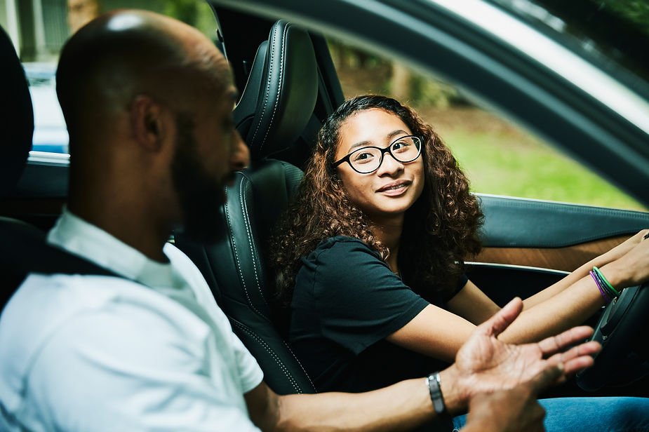 A girl with glasses drives a car while an adult man talks to her. They seem engaged. Car interior is dark with green scenery outside.