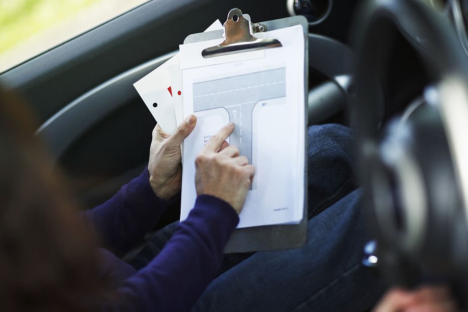 Person in a car points at a diagram on a clipboard, possibly giving directions. The mood is focused, with a blurred steering wheel nearby.