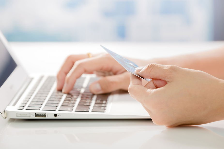 Hands using a laptop and holding a credit card in a bright setting, indicating online shopping. Keyboard keys and blue card visible.