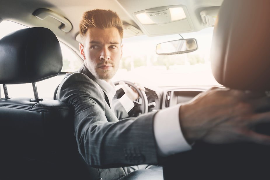 Man in a suit driving a car, looking back over his shoulder with a focused expression. Bright interior lighting and blurred window view.