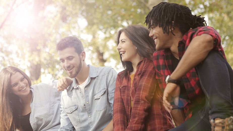 Group of four friends laughing in a sunlit park. Two wearing plaid shirts, two in denim. Green trees in the background, joyful mood.