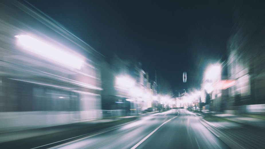 Blurred nighttime street scene with bright lights illuminating buildings on both sides, creating a sense of fast motion and urban energy.