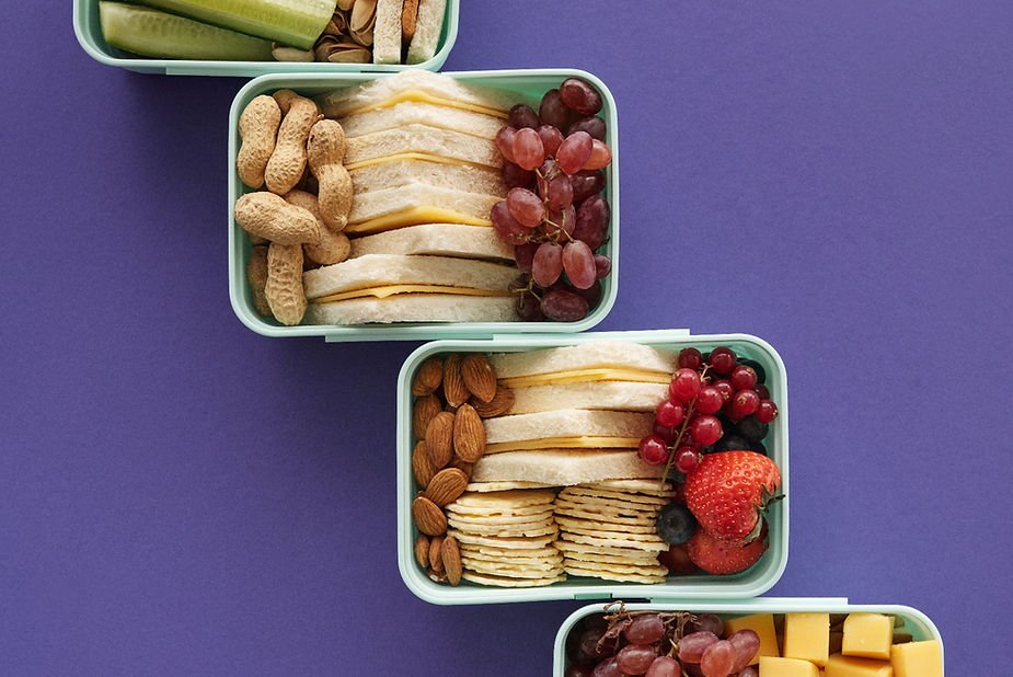 Four lunchboxes with sandwiches, peanuts, almonds, grapes, strawberries, crackers, and cheese on a purple background. Vibrant and organized.