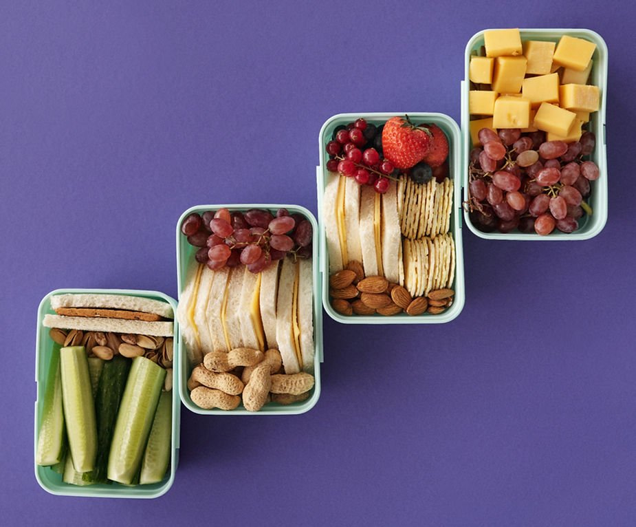 Four lunchboxes on purple background, filled with sandwiches, cubes of cheese, crackers, cucumbers, peanuts, almonds, and fruits like grapes.
