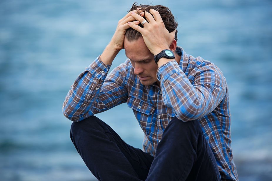 Man in plaid shirt sitting by the water with head in hands, conveying stress. Blue ocean in background, wearing a watch.