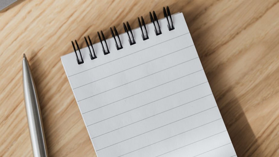 Silver pen beside a lined, spiral notebook on a wooden surface, suggesting a writing or note-taking setting.